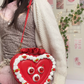 Person holding a red heart-shaped bag with a face design in a room.
