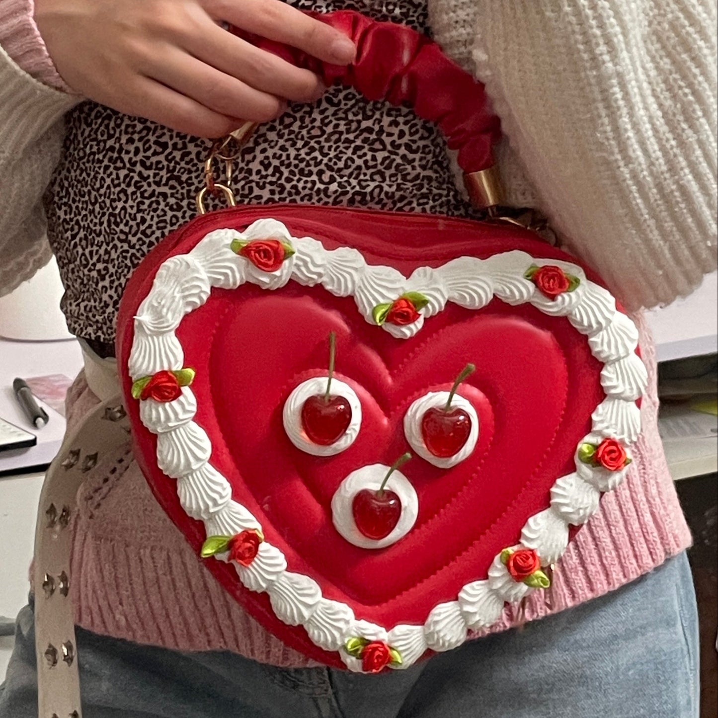 Heart-shaped red bag with white details and cherries held by a person in a casual setting.