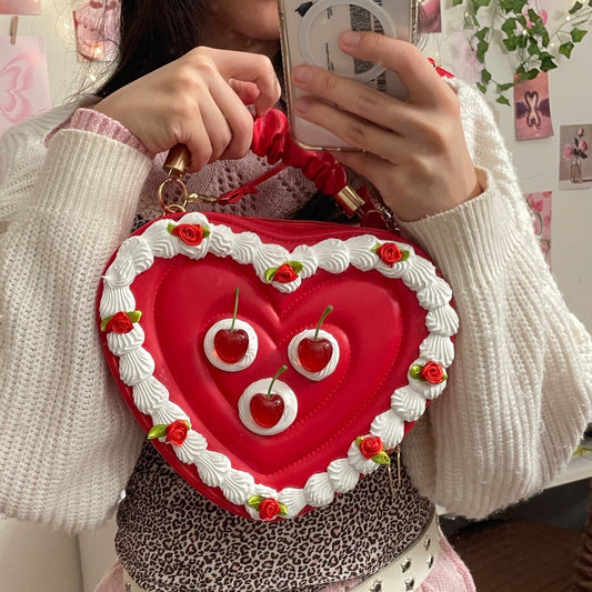 Person holding a heart-shaped cake-themed handbag with cherry decorations.